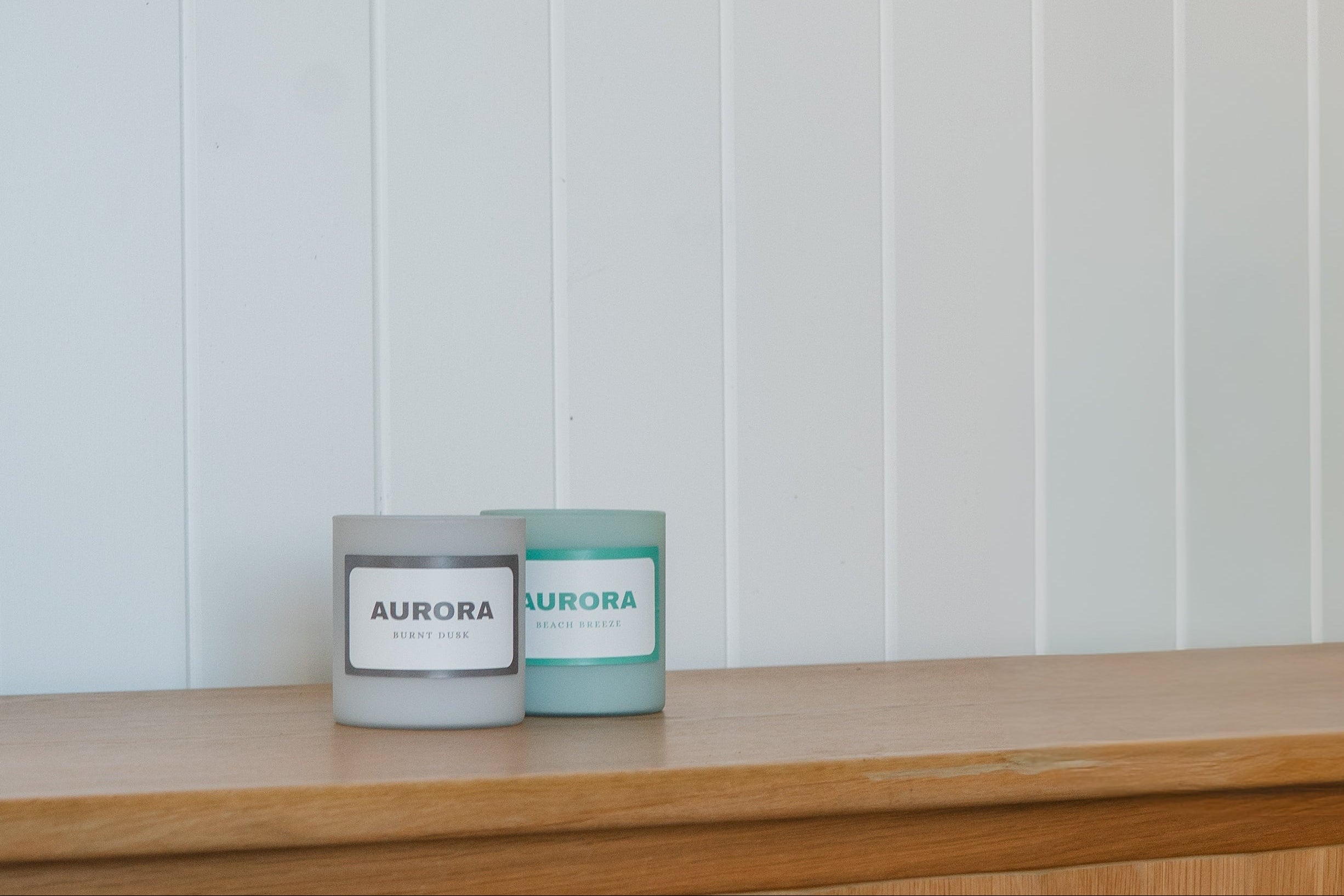 Two small AURORA candles in glass jars sit on a light wood cabinet against a white paneled wall, with a large ocean artwork hanging above.